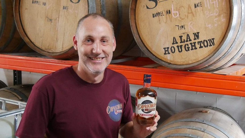 A man sits in front of barrels in a distillery holding a bottle of rum. He is smiling.