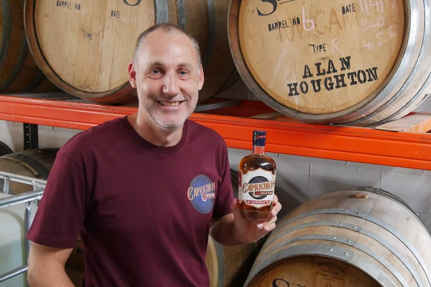 A man sits in front of barrels in a distillery holding a bottle of rum. He is smiling.