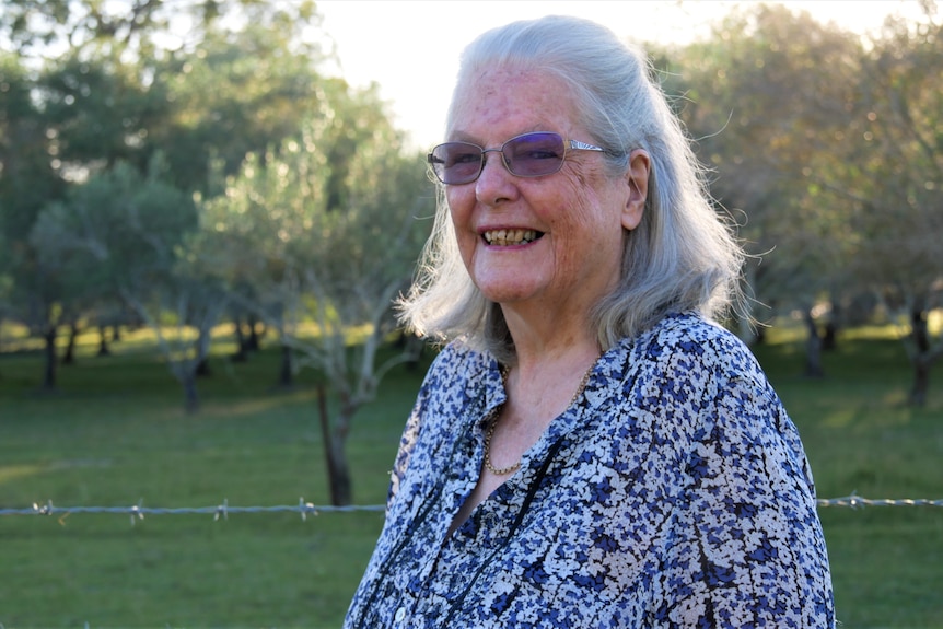 An elderly woman smiles standing in front of a barbed wire fence & olive trees. The setting sun illuminates her hair. 