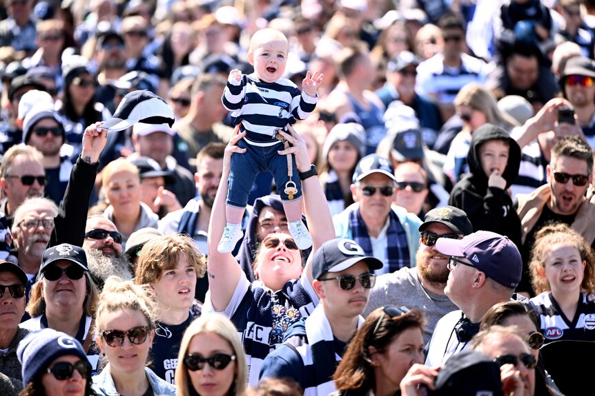 A woman holds a baby up high in a sea of Geelong Cats supporters.