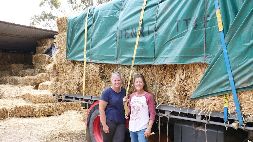 Two women stand in front of a loaded truck filled with hay.