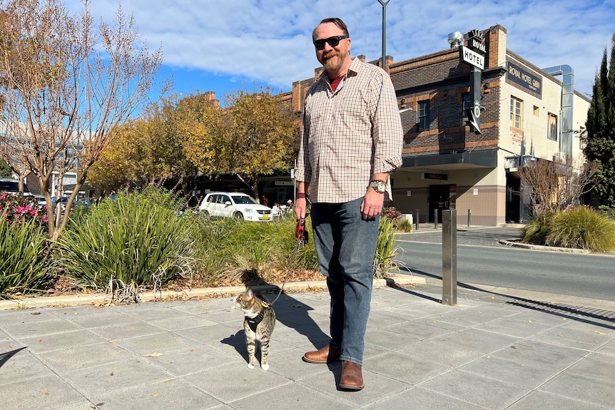 Man wearing a long-sleeved checked shirt walking outside with a cat on a lead. 
