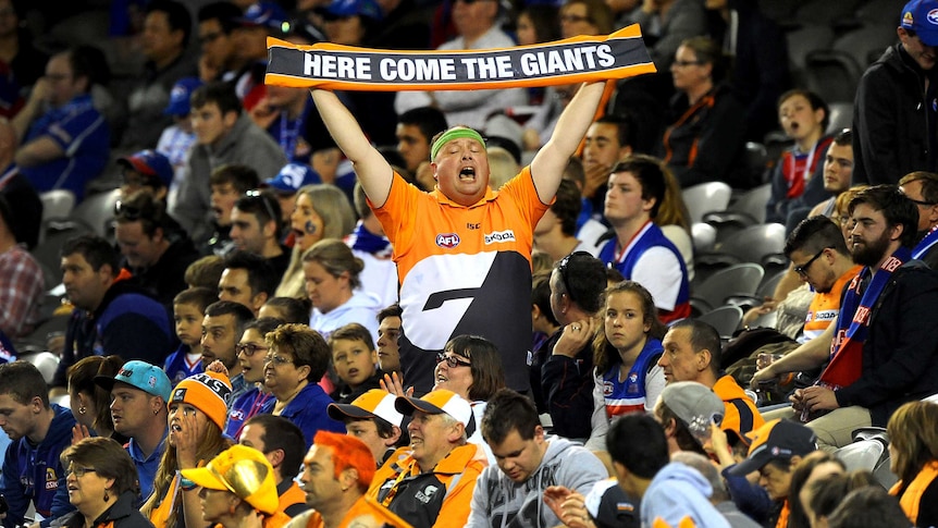 An AFL fan stands waving a banner, surrounded by fans of the opposing team.