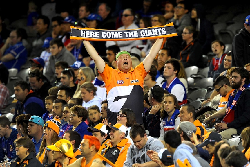 An AFL fan stands waving a banner, surrounded by fans of the opposing team.