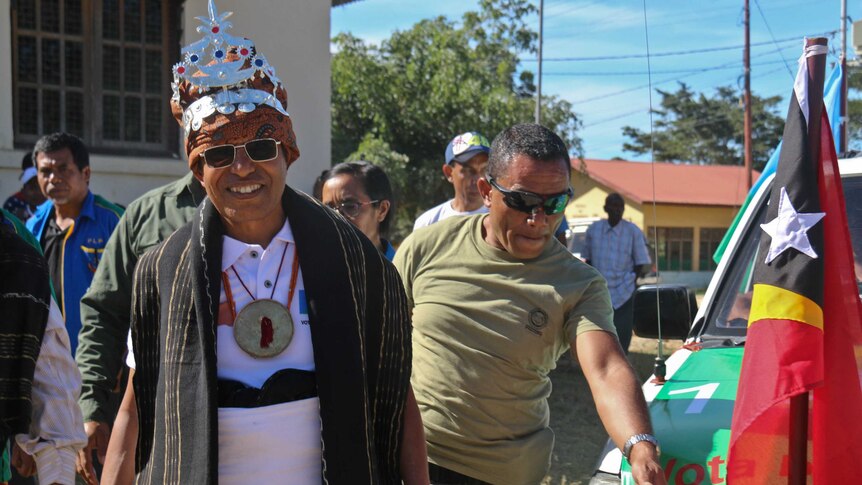 Man wearing silver crown with red and blue jewels walking with a bodyguard near a car.