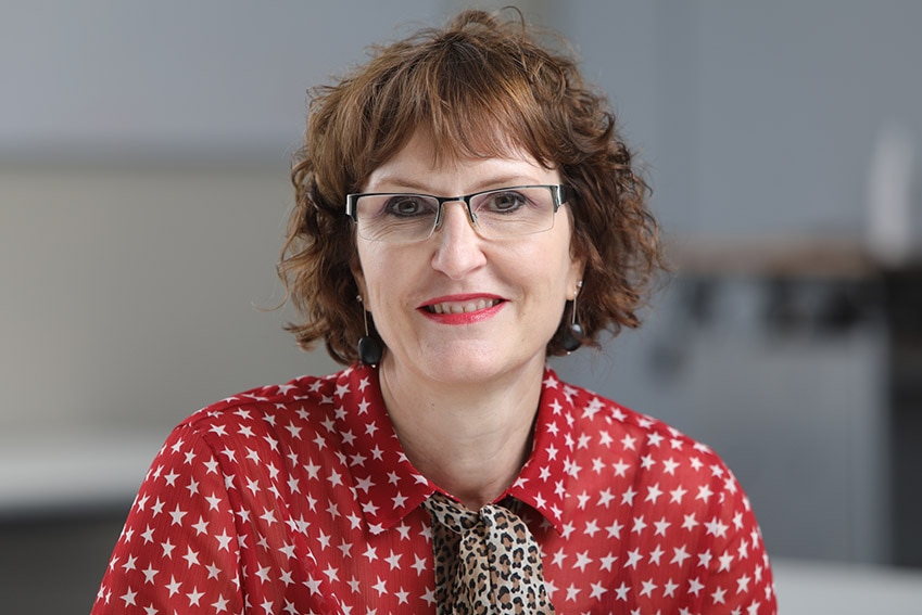 A woman with brown hair and glasses sitting in front of a grey background.