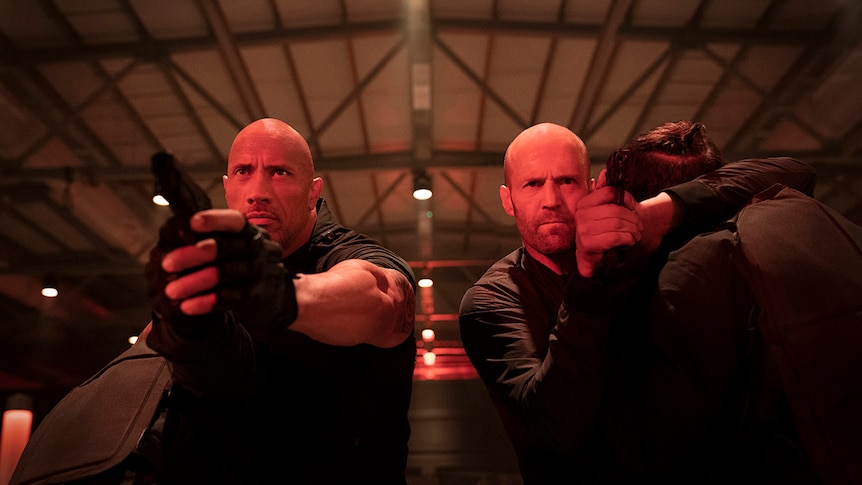 Two men with one arm raised holding handguns in a warehouse with red tinted lighting.