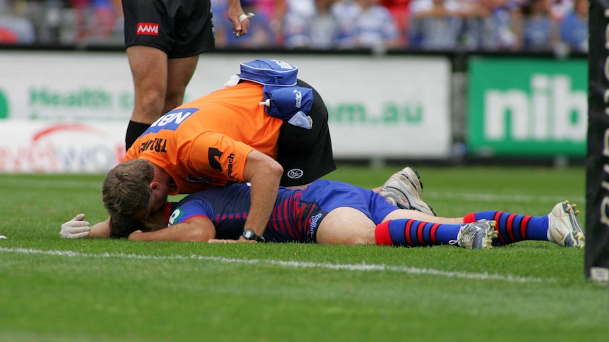 Andrew Johns lies face-down on the grass with a man in orange standing crouched over him