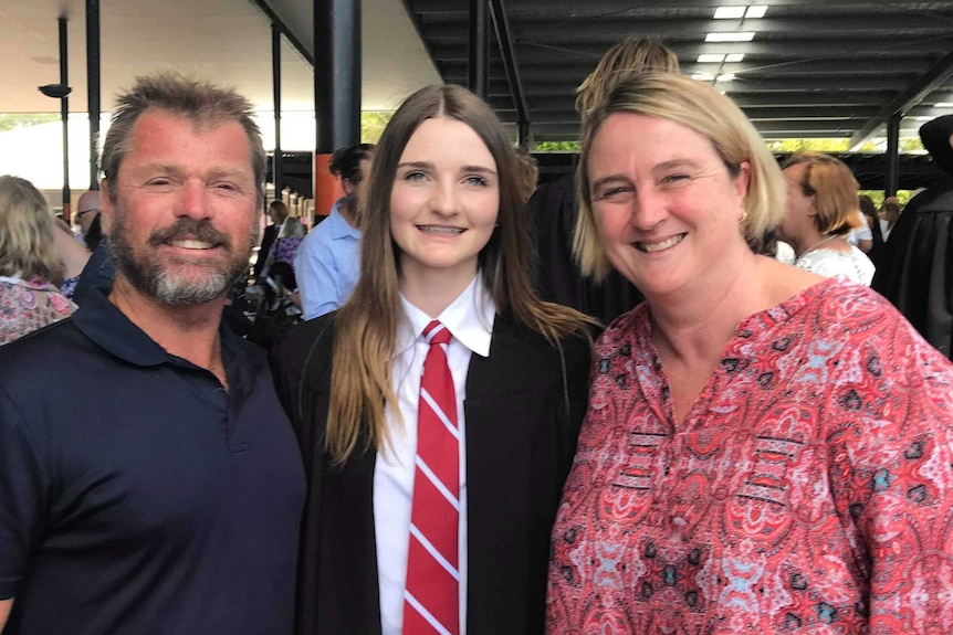 Keely Johnson, centre, in graduation robe with red tie, stands next to her father, left, and mother, right.