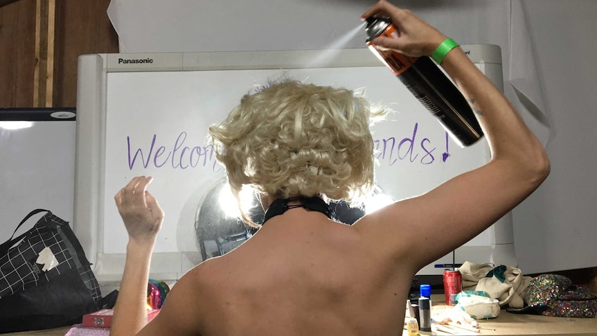 Alicia Zaman uses hairspray on her blonde wig, facing a mirror. Her back is to the camera and the hairspray mist clouds her