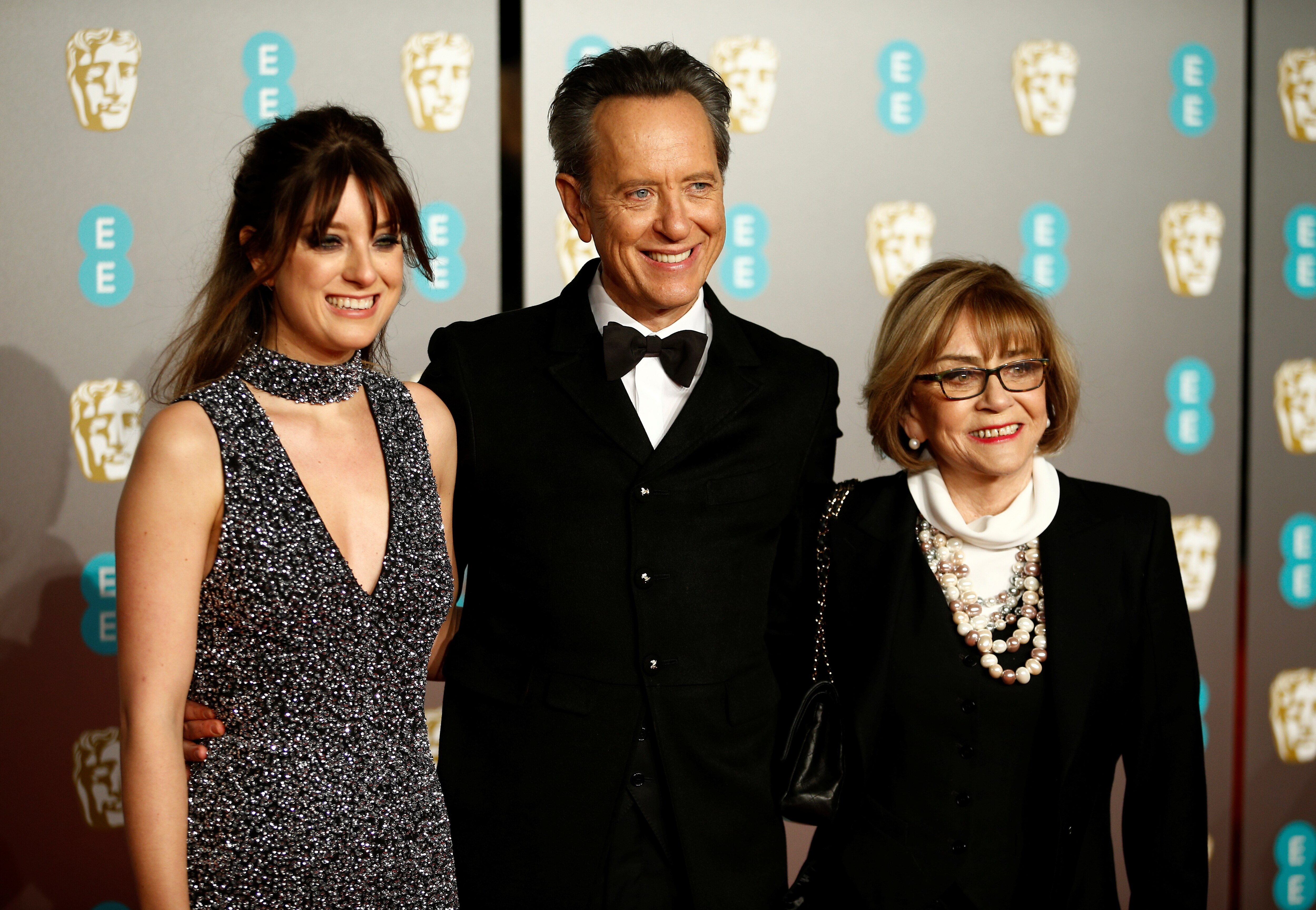 A man in a tuxedo and bow tie is flanked by a young woman in a sparkly dress and an older woman in a black outfit