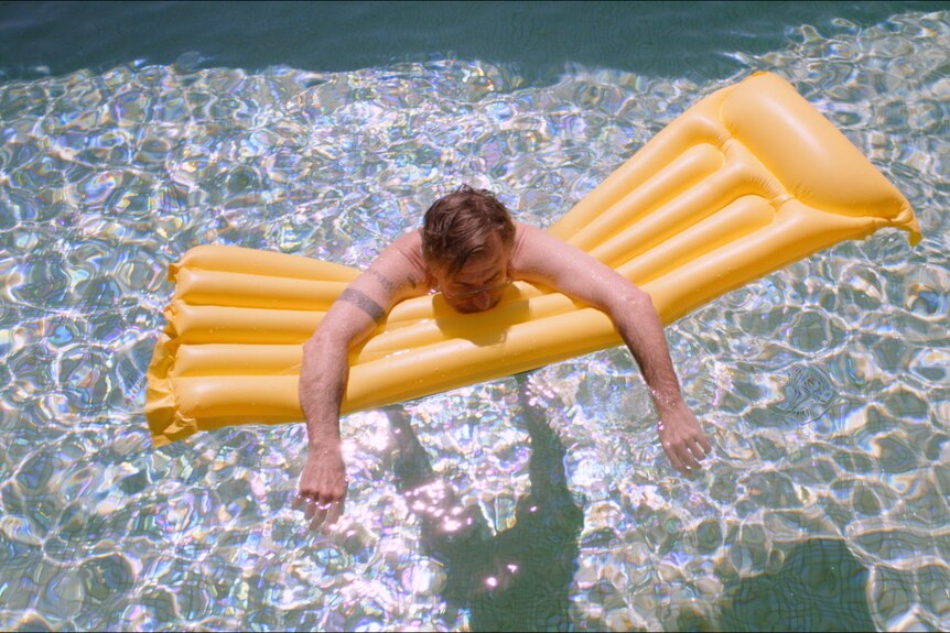 White man with greying beard and short mousey hair floats limply on a yellow inflatable bed in a pool.
