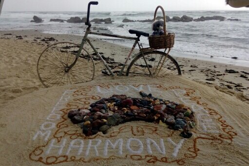 A bicycle with a toy koala in its basket on the sand next to a rock sculpture in the shape of Australia, with 'harmony' under it