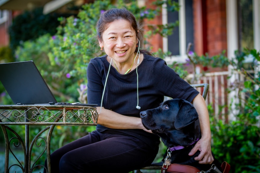 Lee Chong sits smiling at a small metal table outdoors, with her arms around her black guide dog Genie.