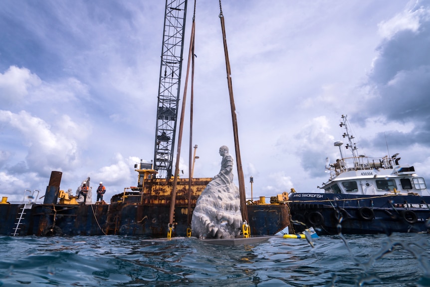 A large concrete statue is lowered off a ship into the ocean by a crane