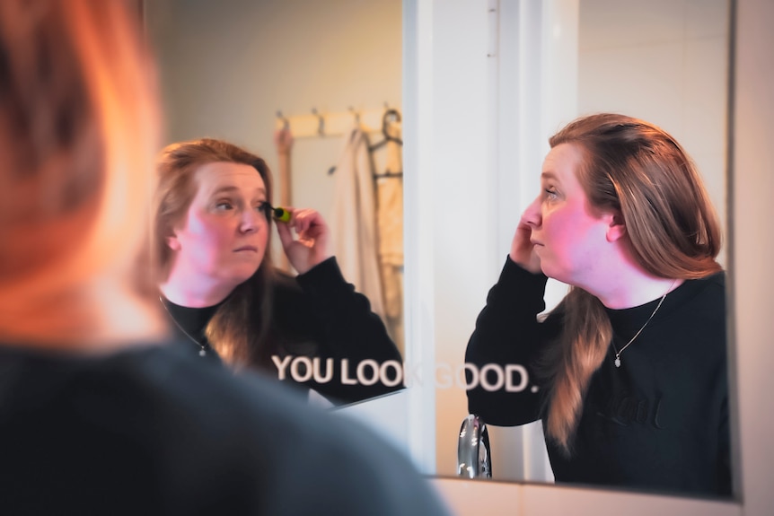 31-year-old Jasmine puts on mascara in the bathroom mirror. On the mirror are the words 'You Look Good'.