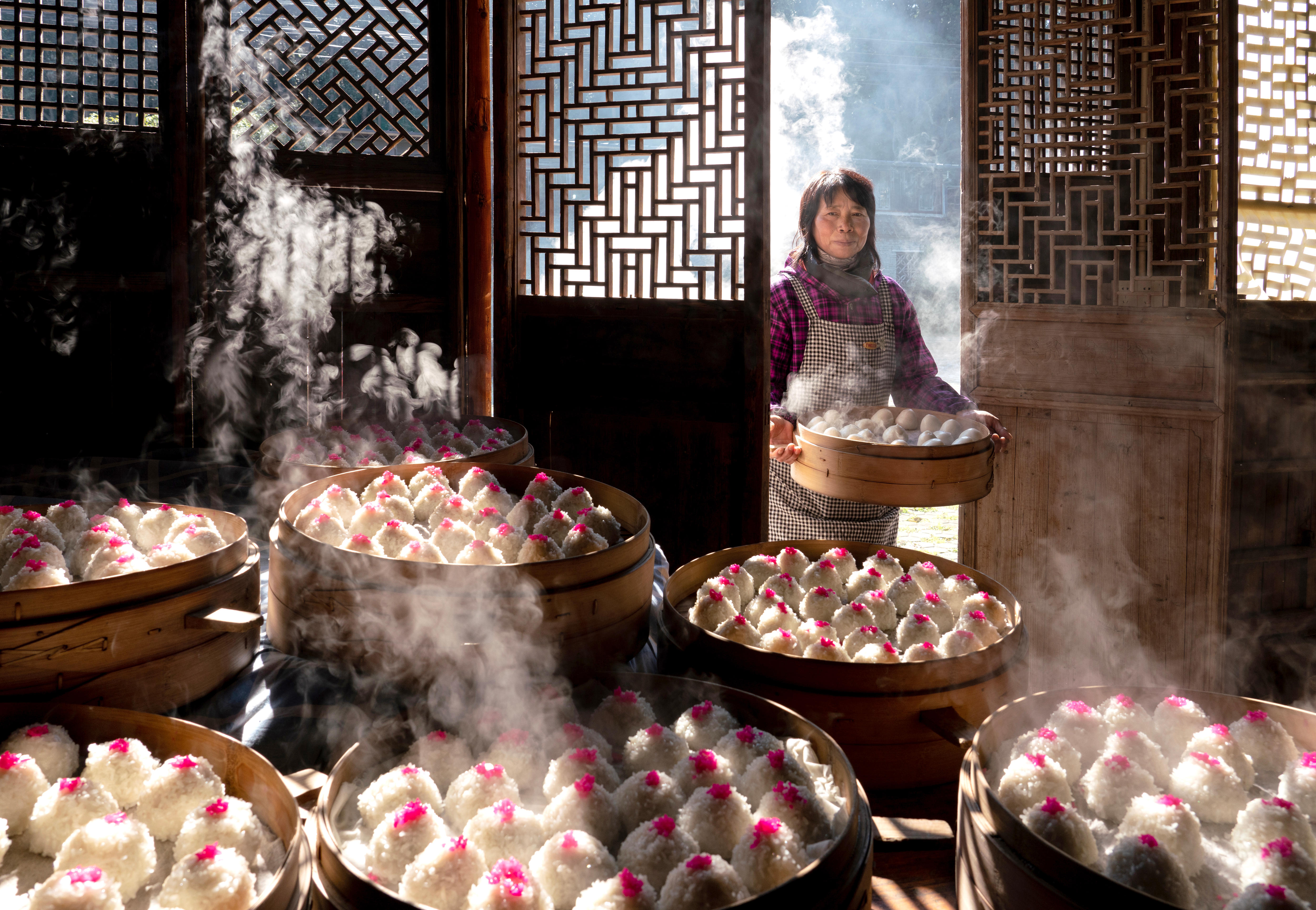 A woman carries a tray of red bean paste balls through a door. Inside are more trays, holding hundreds of balls with steam.