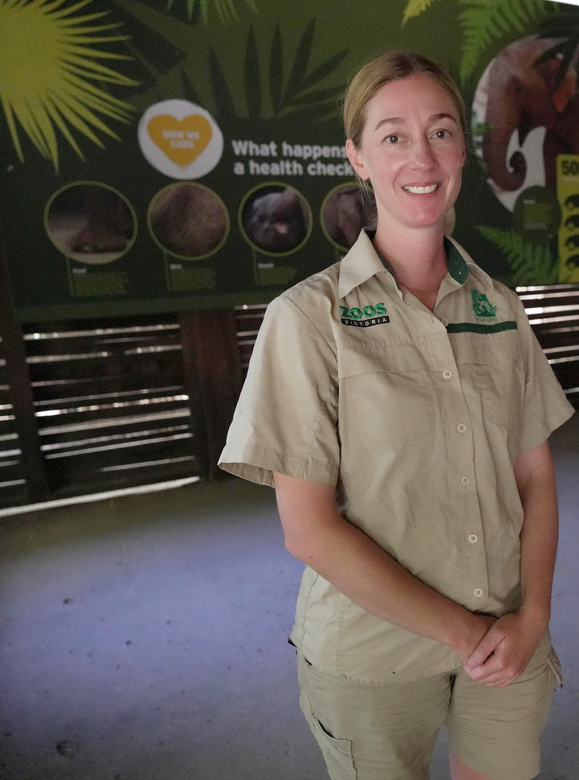 A woman in a beige short-sleeve shirt with zoo logos standing in front of an elephant display.