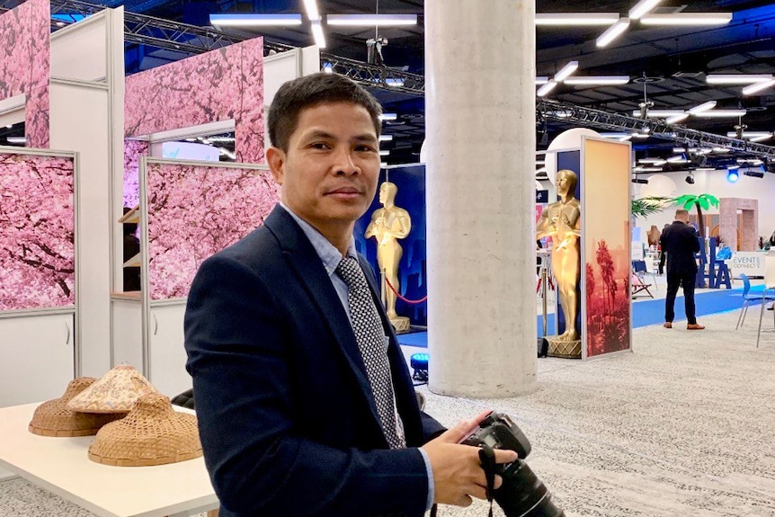 a man wearing a suit stands in a large conference hall with artworks behind him.