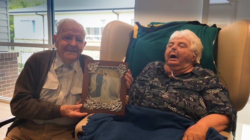 An elderly man and wife, the man holding a black and white wedding photo, she in a chair with tubes in her nose