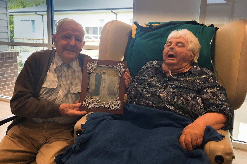 An elderly man and wife, the man holding a black and white wedding photo, she in a chair with tubes in her nose