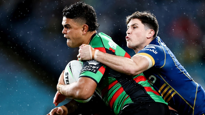 A South Sydney NRL player holds the ball as he is tackled by Parramatta opponents.