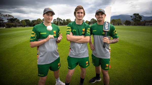 Three young men in cricket gear, one holds a ball, another a bat
