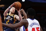Joe Ingles grimaces and holds the basketball outstretched as a two defenders close in on him