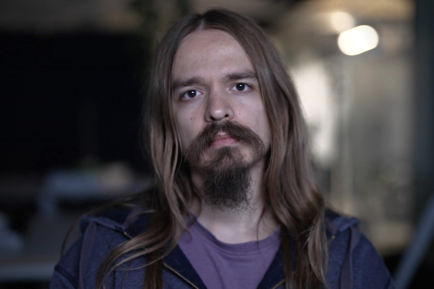 Man with long hair and a beard in a purple shirt and blue hoodie.