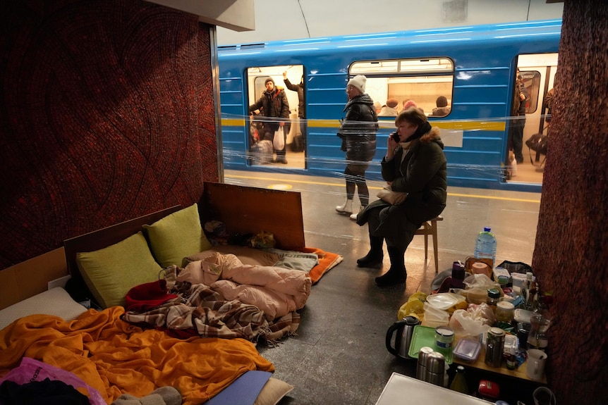 Woman sits in improvised shelter in Kyiv metro station as train stops behind.