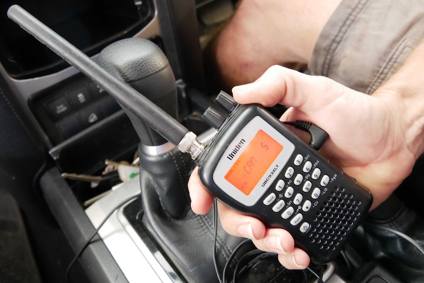 Males hand holding black hand held radio that is plugged into the centre console of a car