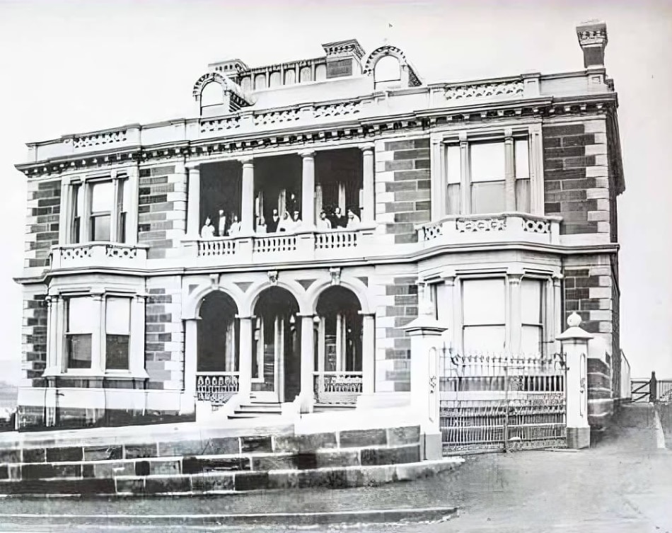 Una fotografía en blanco y negro de un ornamentado edificio de dos pisos con porches y gente en el último piso.