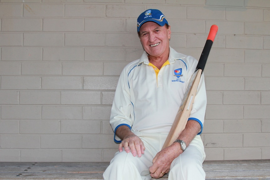 Elderly cricketer sits on the bench wearing his white cricket uniform and holding a cricket bat.