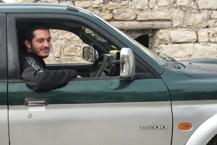 Man with beard wearing tracksuit jumper smiles sitting in car looking out window with small stone building in background.