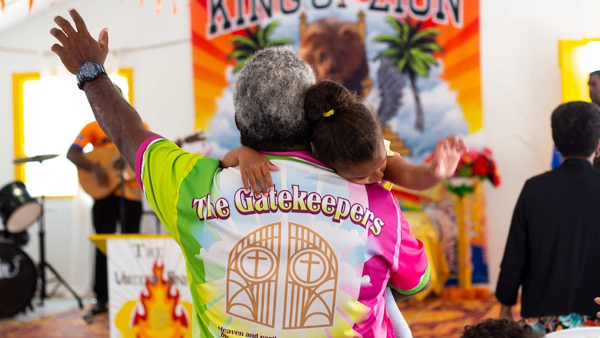 A man holds up a hand, holding a young girl, inside a church with a sign that reads, 'King of Zion'.