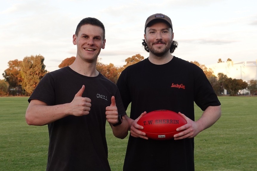 Two students stand next to each other on football oval, one giving the thumbs up