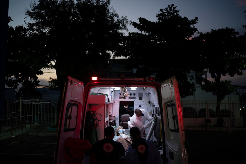 The back of an ambulance is opened to reveal a masked patient surrounded by medical staff at dusk.