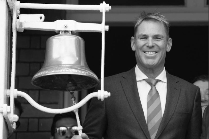 Black and white image of smiling Shane Warne in suit and tie ringing a bell