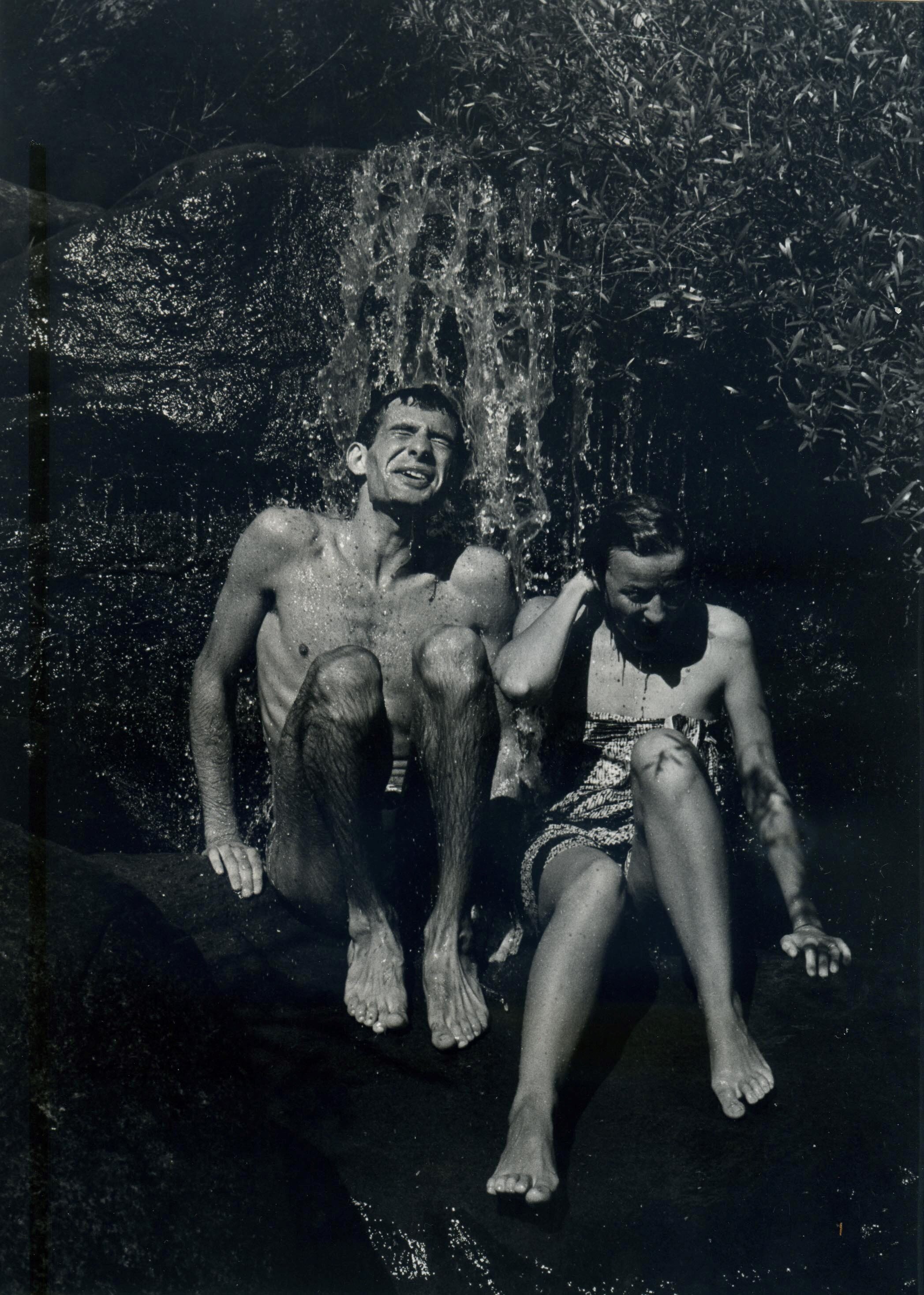 A black and white photo of a man and a women wearing swimmers sitting under a waterfall