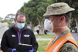 A man in an army uniform and fluorescent vest stands on a suburban footpath with a man with a clipboard.