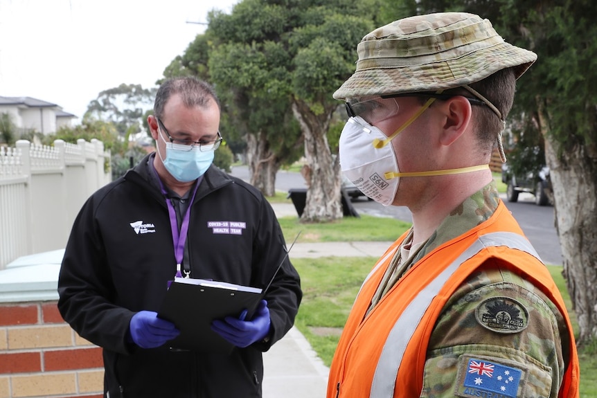 A man in an army uniform and fluorescent vest stands on a suburban footpath with a man with a clipboard.