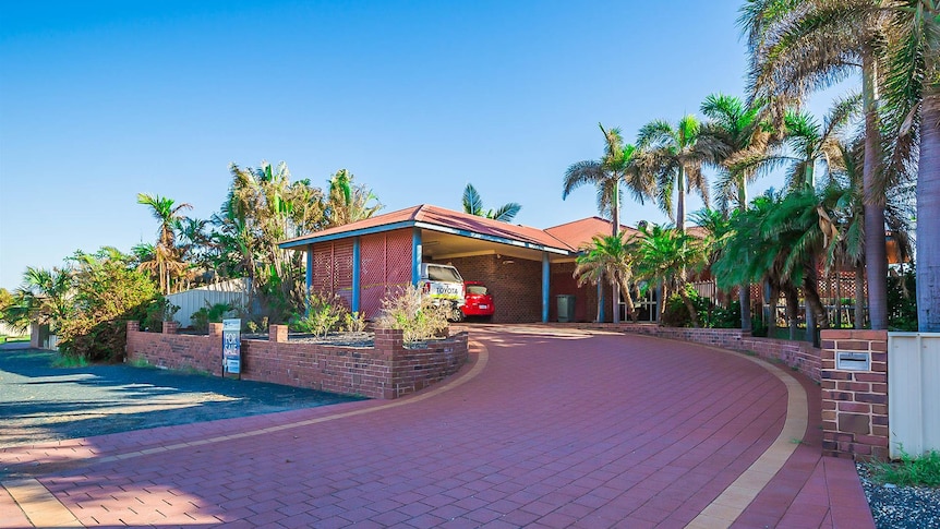 A red brick house with a red brick driveway with lots of tall palm trees surrounding it.
