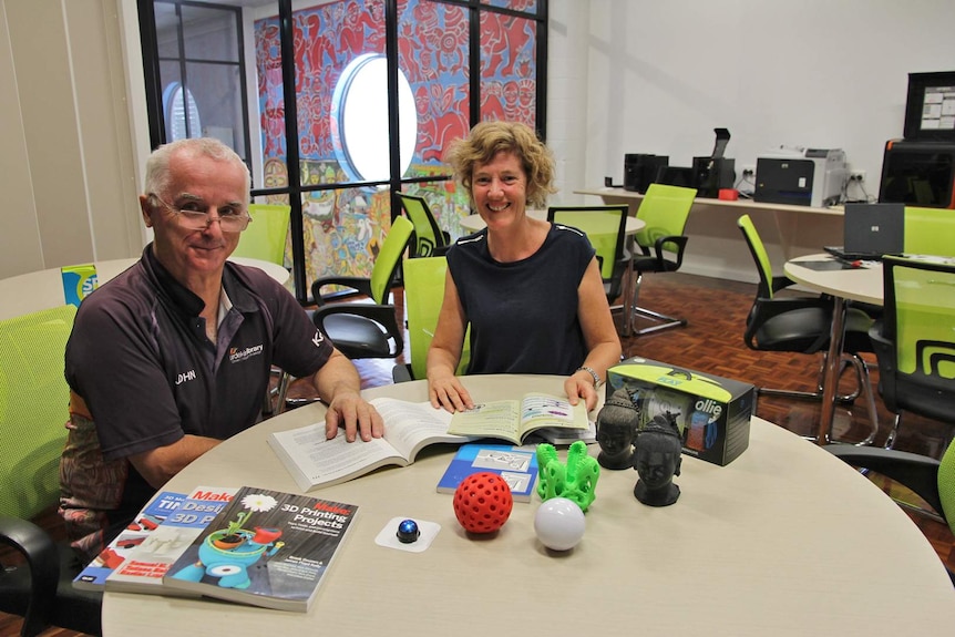 A man and woman sit at a round table with 3D printed sculptures and books about 3D printing on the desk
