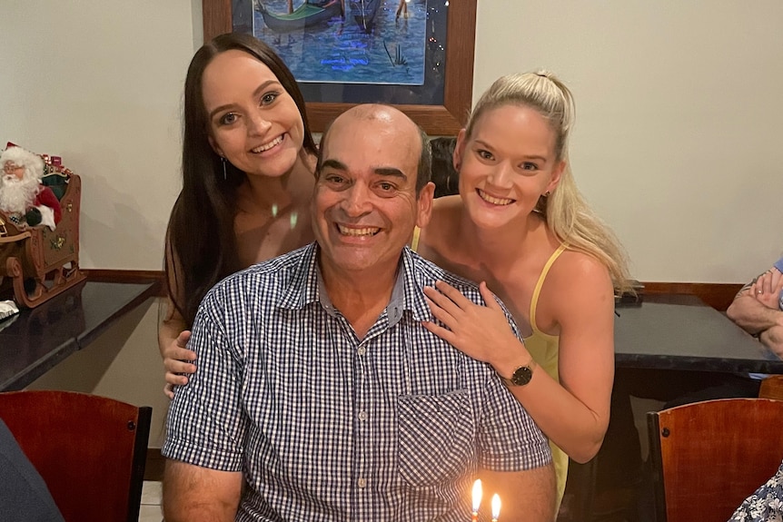 Paul Muscat seated in between his two daughters Emma (left) and Kate (right), both daughters have an arm around him