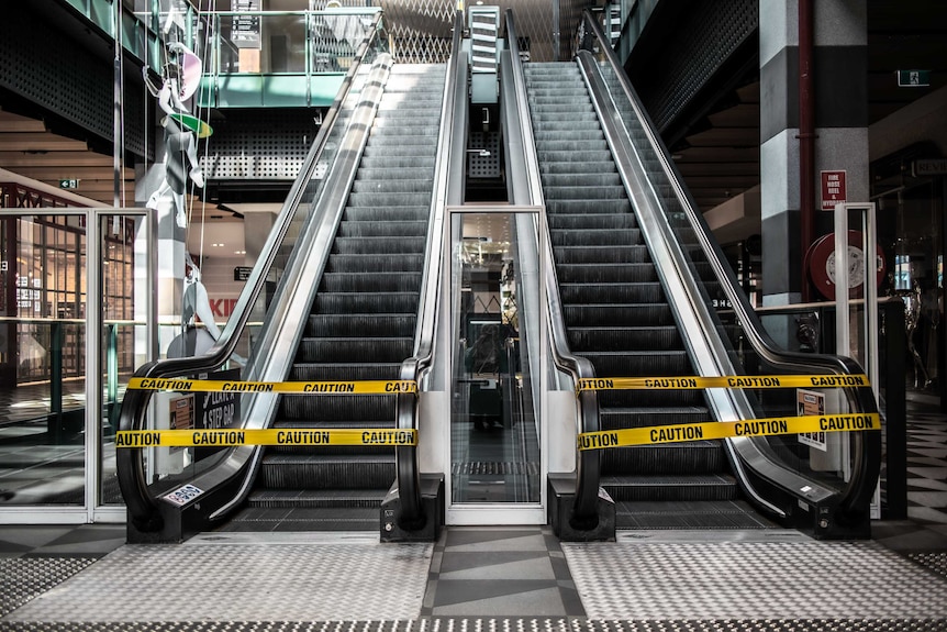 Two escalators are blocked by yellow tape in an empty shopping centre.