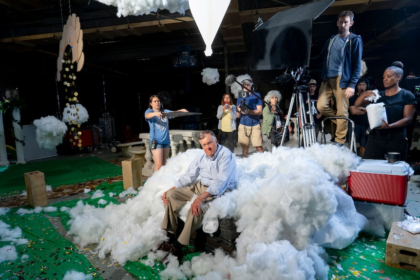An older man in khaki slacks and blue shirt sits on couch surrounded by cotton on green fabric, behind him are film set crew.