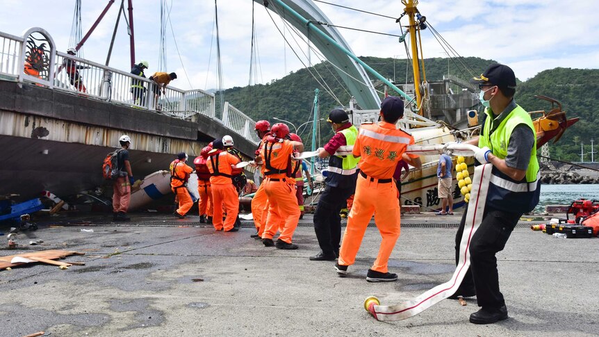 At least 10 people pull a rope as the majority wear orange reflective gear. A bridge is above them.