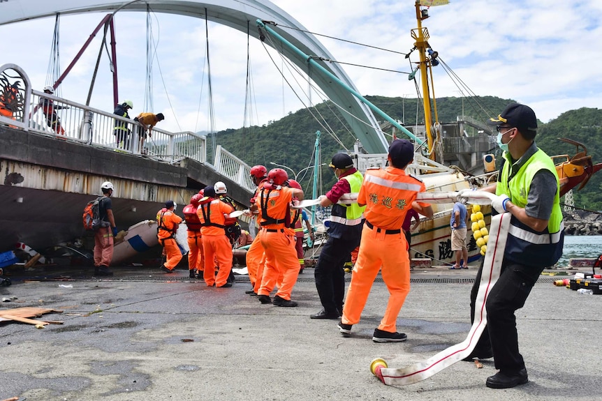 At least 10 people pull a rope as the majority wear orange reflective gear. A bridge is above them.