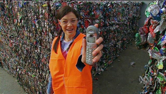 A woman holds up a mobile phone while standing in front of bale of compressed aluminium cans