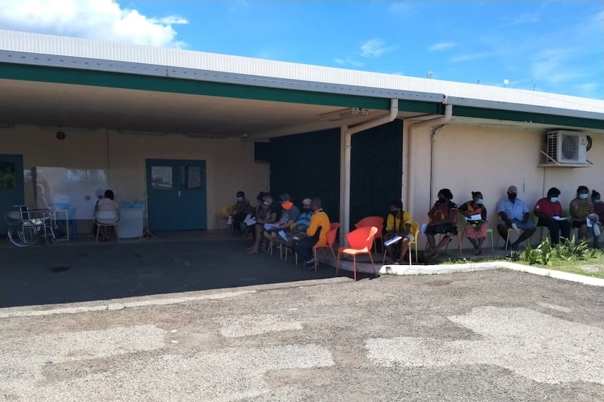 People sit in chairs in the a queue in the shade around a one story building.